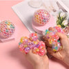 A hand holds a vibrant, multi-colored rainbow squishy stress relief ball against a pink background.