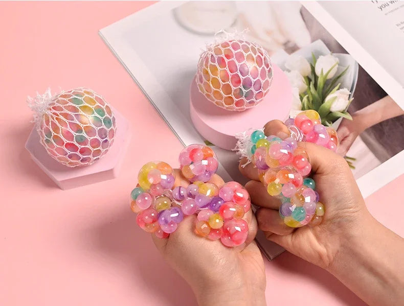 A hand holds a vibrant, multi-colored rainbow squishy stress relief ball against a pink background.