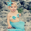 A baby wearing a turquoise knit tail skirt and a matching pearl headband posing outdoors.