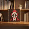 A square pixel art board featuring a red and white stylized face design, displayed on a wooden shelf among books.