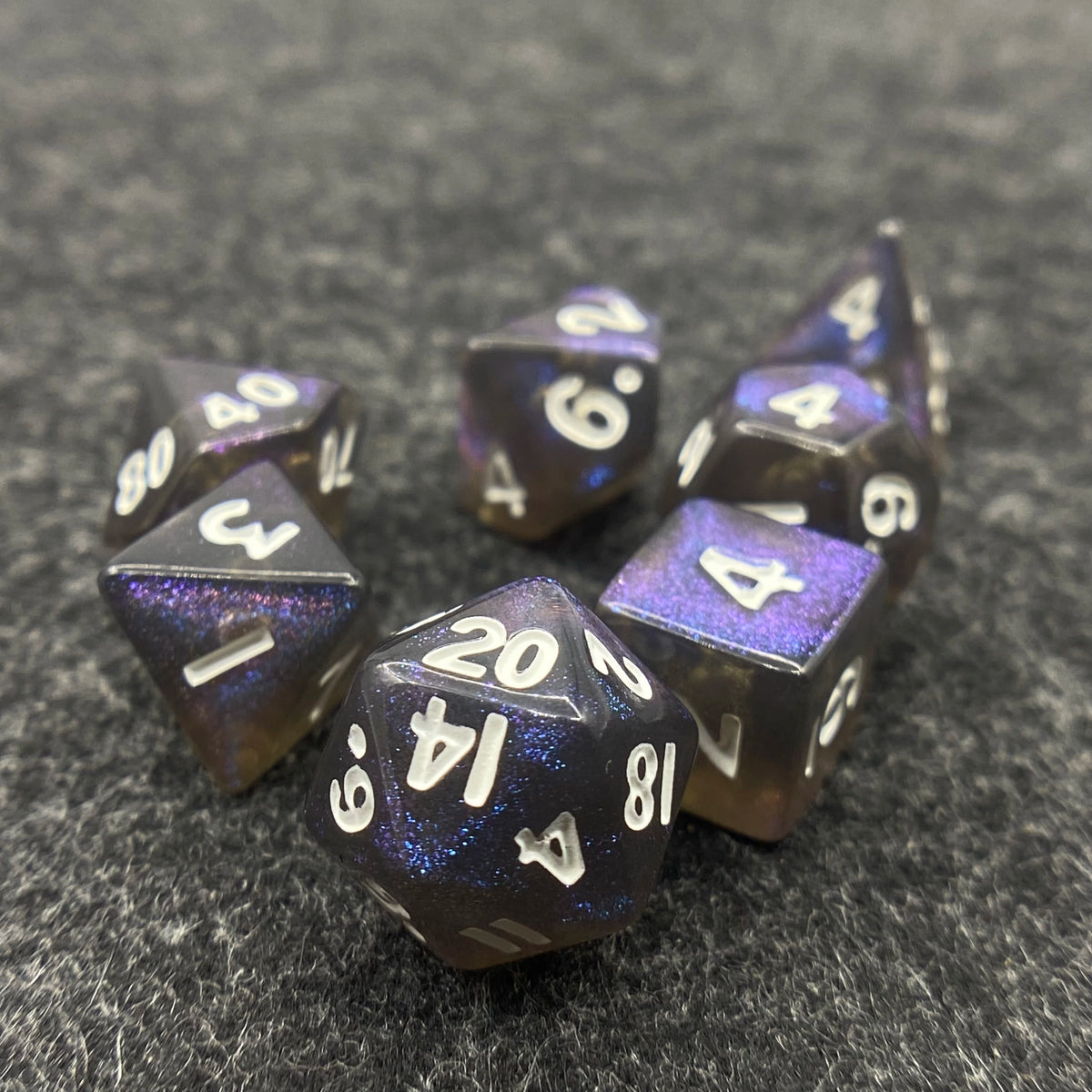 A collection of seven iridescent acrylic polyhedral dice scattered on a dark gray surface.