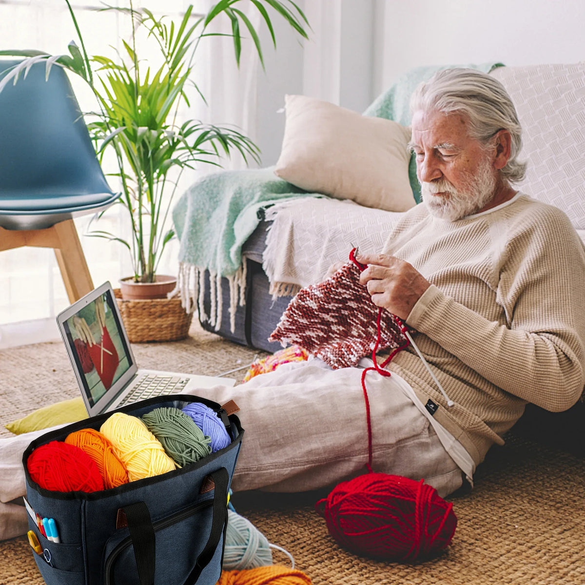 An elderly man sits on the floor surrounded by various yarn and knitting supplies, including a canvas tote bag.