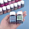 A hand holds two glass bottles of Acid Blue and Grass Green textile fabric dye powder for clothing refurbishment.