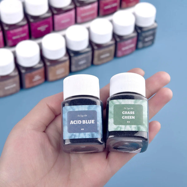 Mano sostiene dos botellas de vidrio con polvo de tinte textil Acid Blue y Grass Green para renovar ropa