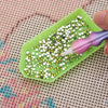 A hand holds a green plastic tray filled with small gold and green sequins, demonstrating a precision diamond painting pen.