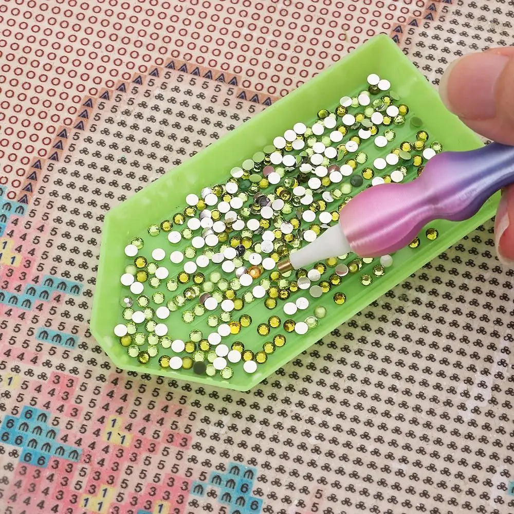 A hand holds a green plastic tray filled with small gold and green sequins, demonstrating a precision diamond painting pen.