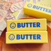 Two yellow squishy toys labeled BUTTER with smiling faces are displayed on a wooden plate.