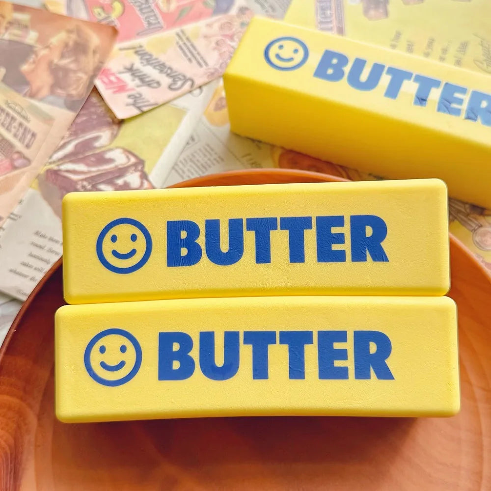 Two yellow squishy toys labeled BUTTER with smiling faces are displayed on a wooden plate.