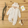 A cream-colored, quilted hooded baby romper lies on a wooden surface next to a sprig of greenery and a yellow smiley face car