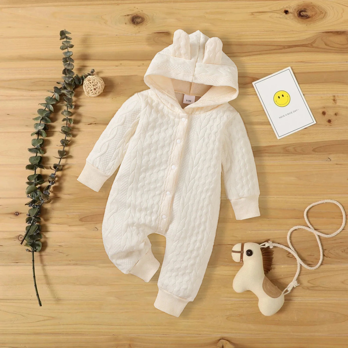 A cream-colored, quilted hooded baby romper lies on a wooden surface next to a sprig of greenery and a yellow smiley face car