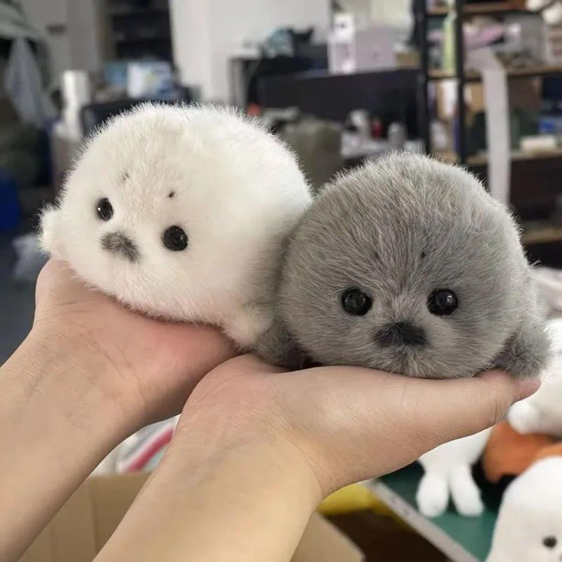 A pair of adorable grey and white seal plush keychains held in a person's hand.