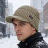 A man wearing a beige knit beanie with a brim and a black jacket in a snowy outdoor setting.