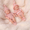 A realistic soft vinyl baby doll wearing a pink striped outfit and matching hat, lying on a fluffy white surface.
