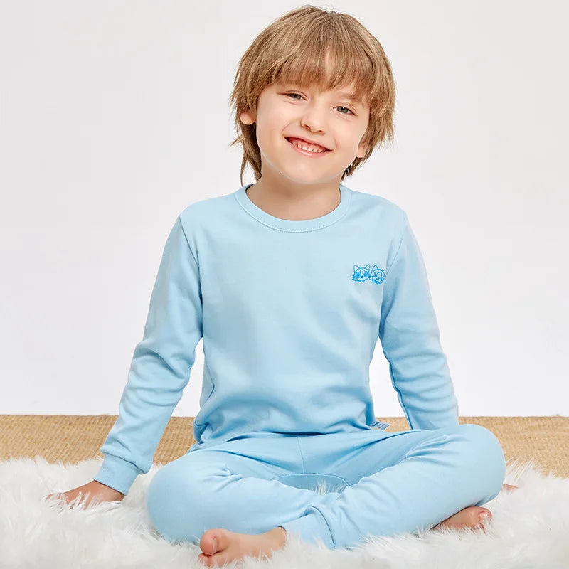 A smiling young boy wearing a light blue cotton pajama set with an astronaut print, sitting on a white fluffy surface.
