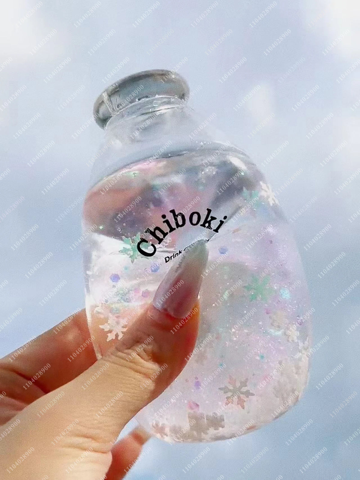 A hand holds a clear plastic bottle filled with liquid and glitter, labeled Chiboki, against a light blue sky.