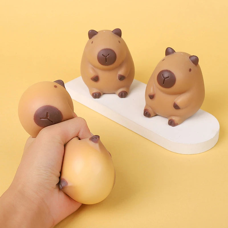 A hand is squeezing a small, round, brown capybara-shaped stress relief toy against a white surface.