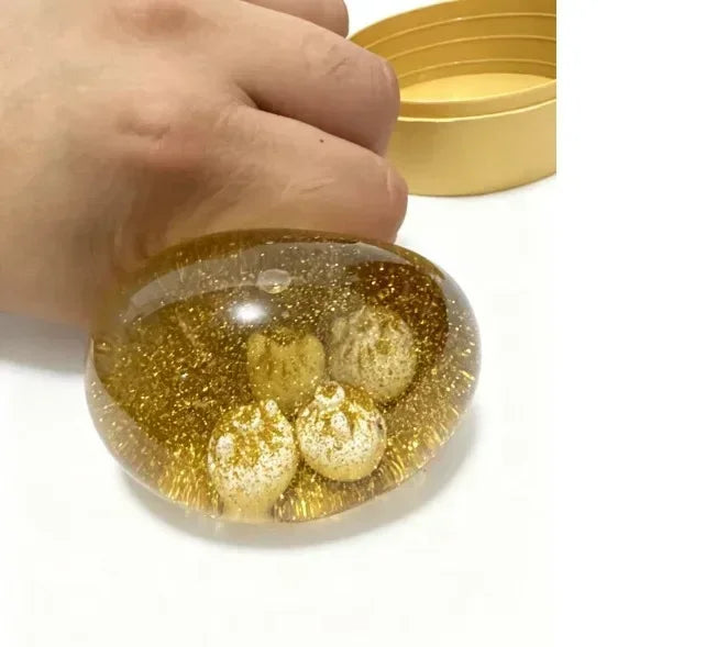 Une main tient un objet en verre rond et transparent rempli de paillettes dorées et de petites billes texturées.