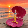 A pink seashell shaped bath toy featuring a mermaid figure with red hair and yellow tail set against a sunset beach backgroun