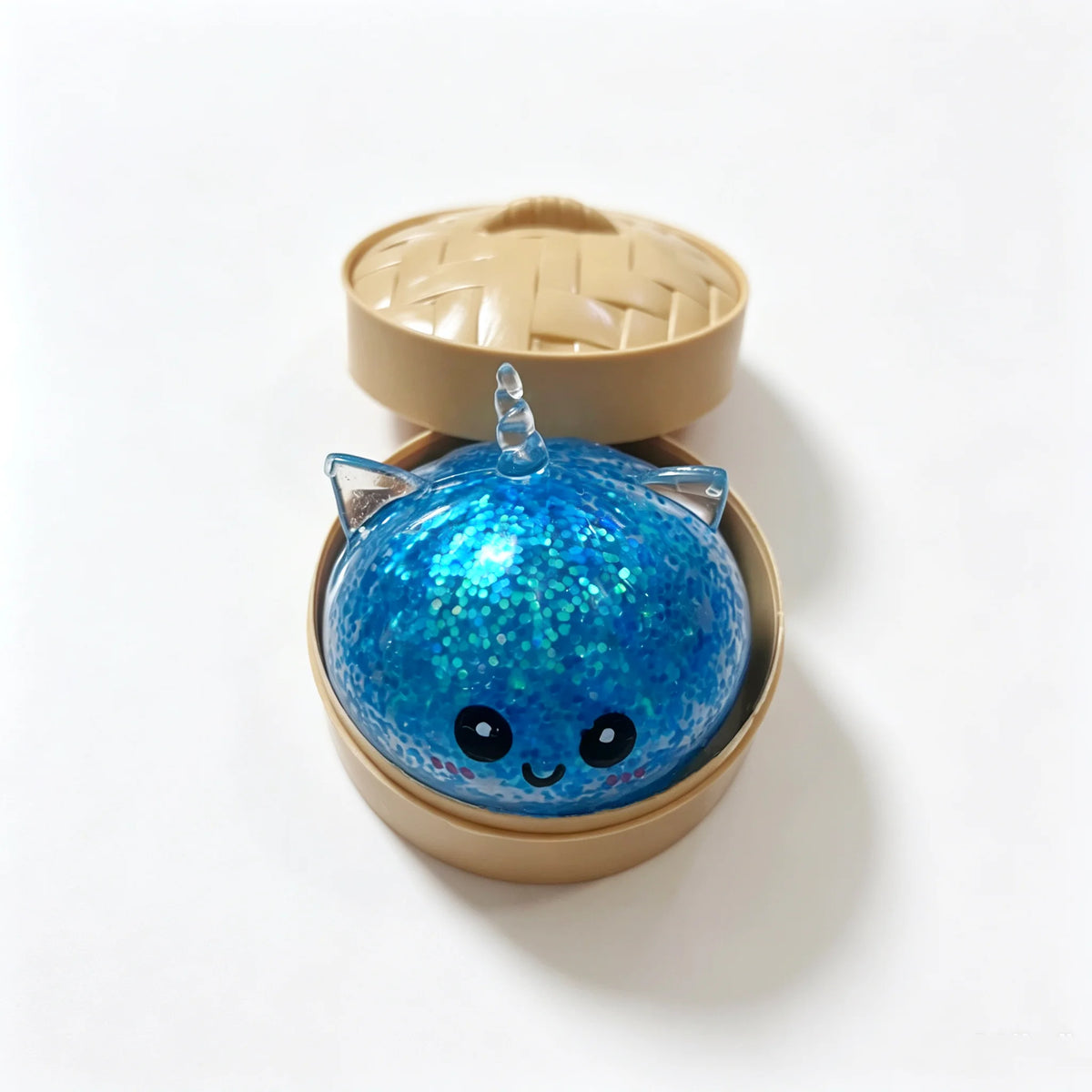A blue glittery unicorn dumpling squishy stress ball resting in a light brown bamboo steamer box.
