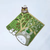 A plush toy of a cat with yellow eyes and green fur is attached to a felt leaf shape featuring a tree and small white animal 