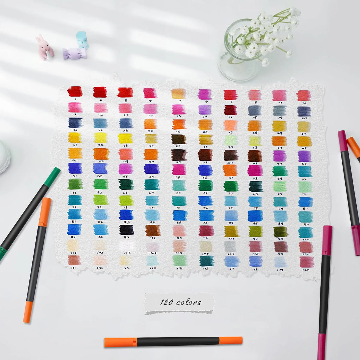A sheet of white paper featuring 120 colored art markers arranged in rows, with several paintbrushes and a small glass of whi