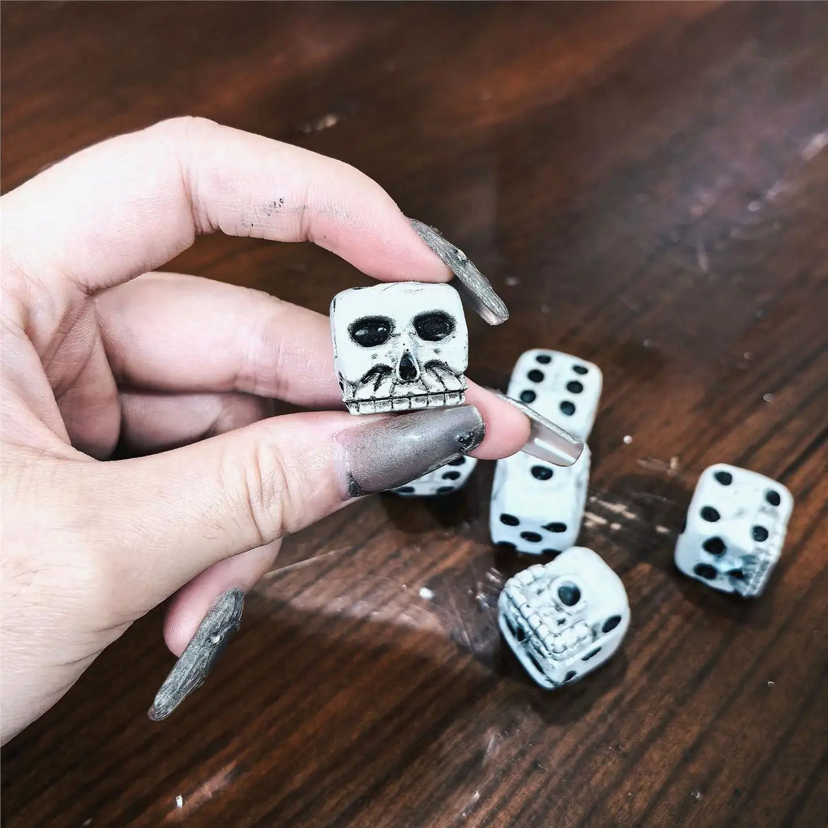 A hand holds a mini white skull dice set for Halloween and party games on a dark wooden surface.