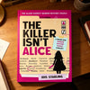 A book titled The Killer Isn't Alice featuring a pink cover and various puzzle elements is displayed on a wooden table.