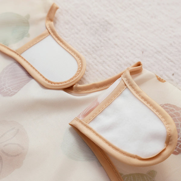 A light beige waterproof long sleeve feeding bib with a white insert and tan straps is shown against a pale patterned fabric.