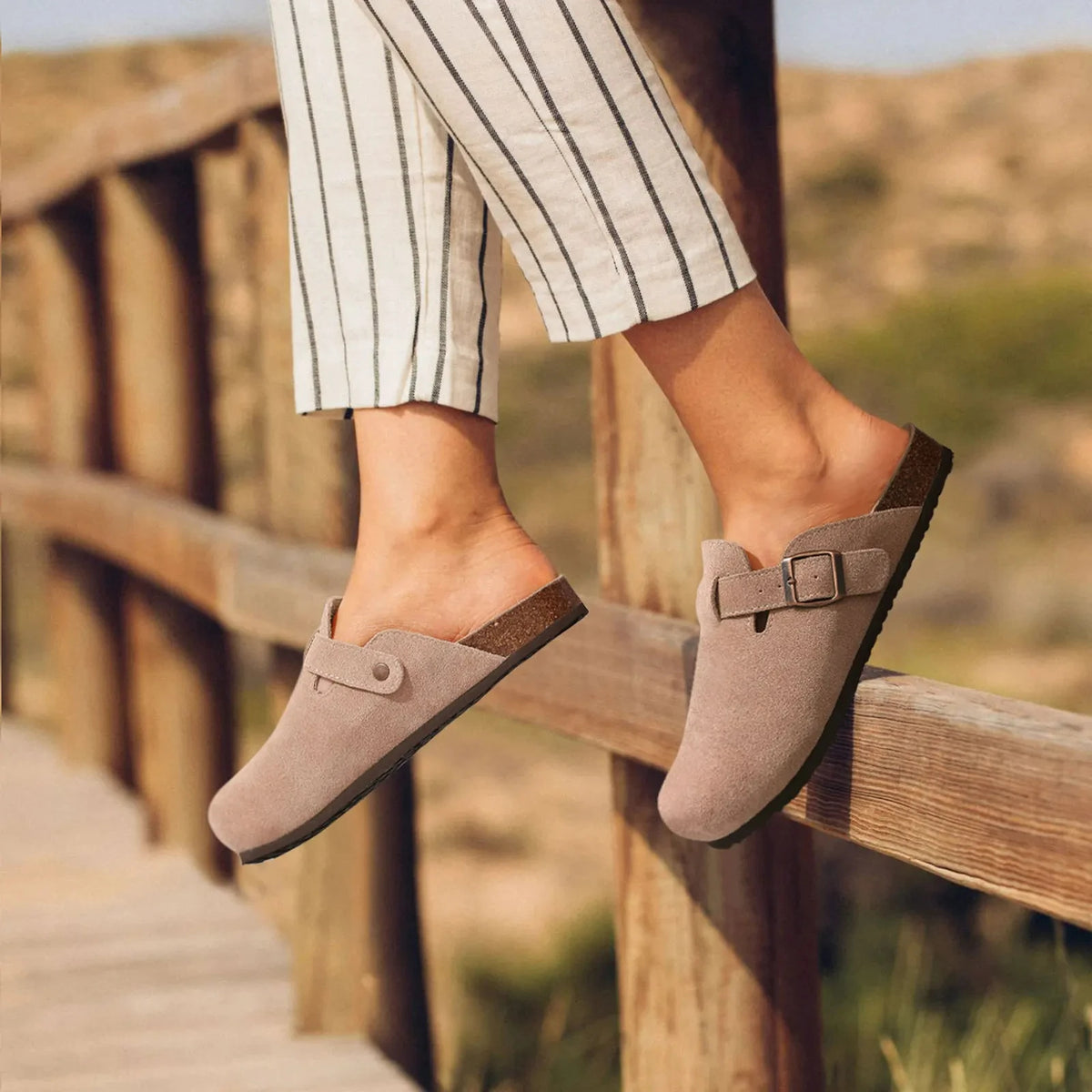 A person wearing taupe suede cork mules with arch support standing outdoors on a wooden deck.