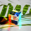 A transparent optical color cube prism with various colored facets rests on a surface amidst green leaves.