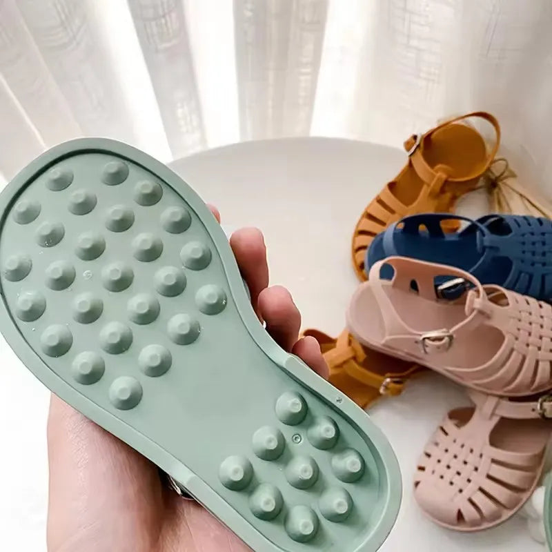 A hand holds a green non-slip sole for girls' gladiator jelly sandals alongside several pairs of similar colored sandals.