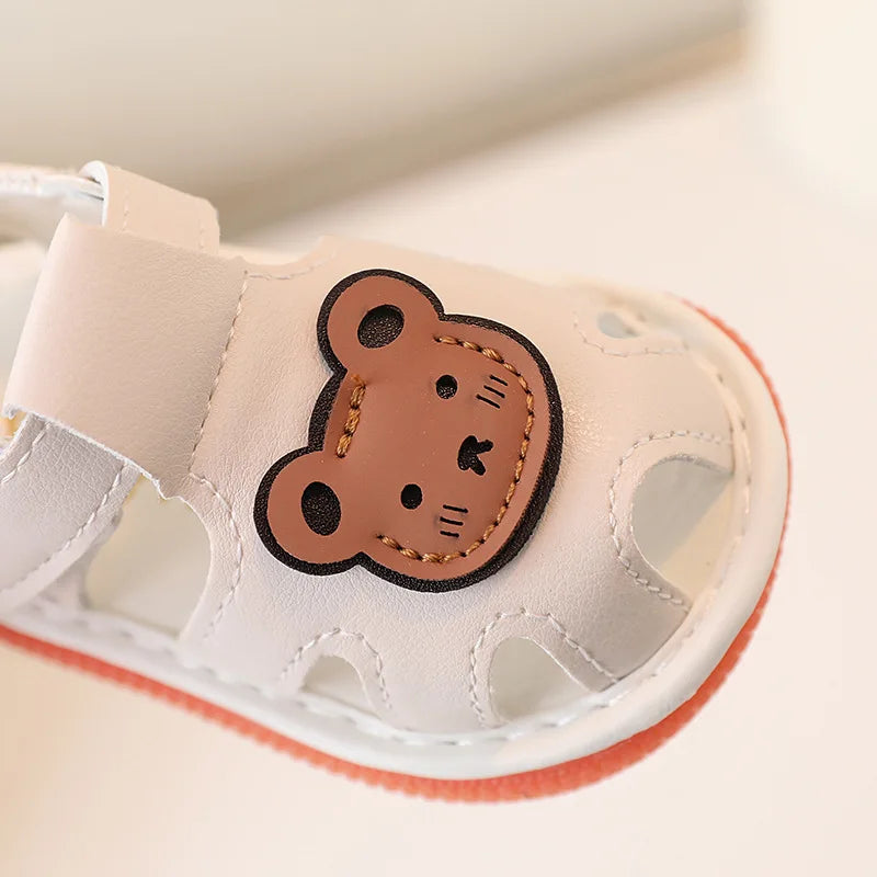 A pair of light beige soft sole sandals for infants and toddlers featuring a cute brown bear design.