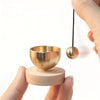 A hand holds a wooden bowl and a hanging brass singing bowl with a dark cord.