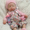 A realistic reborn baby doll wearing a pink knitted hat and floral outfit, lying on a fluffy white surface.