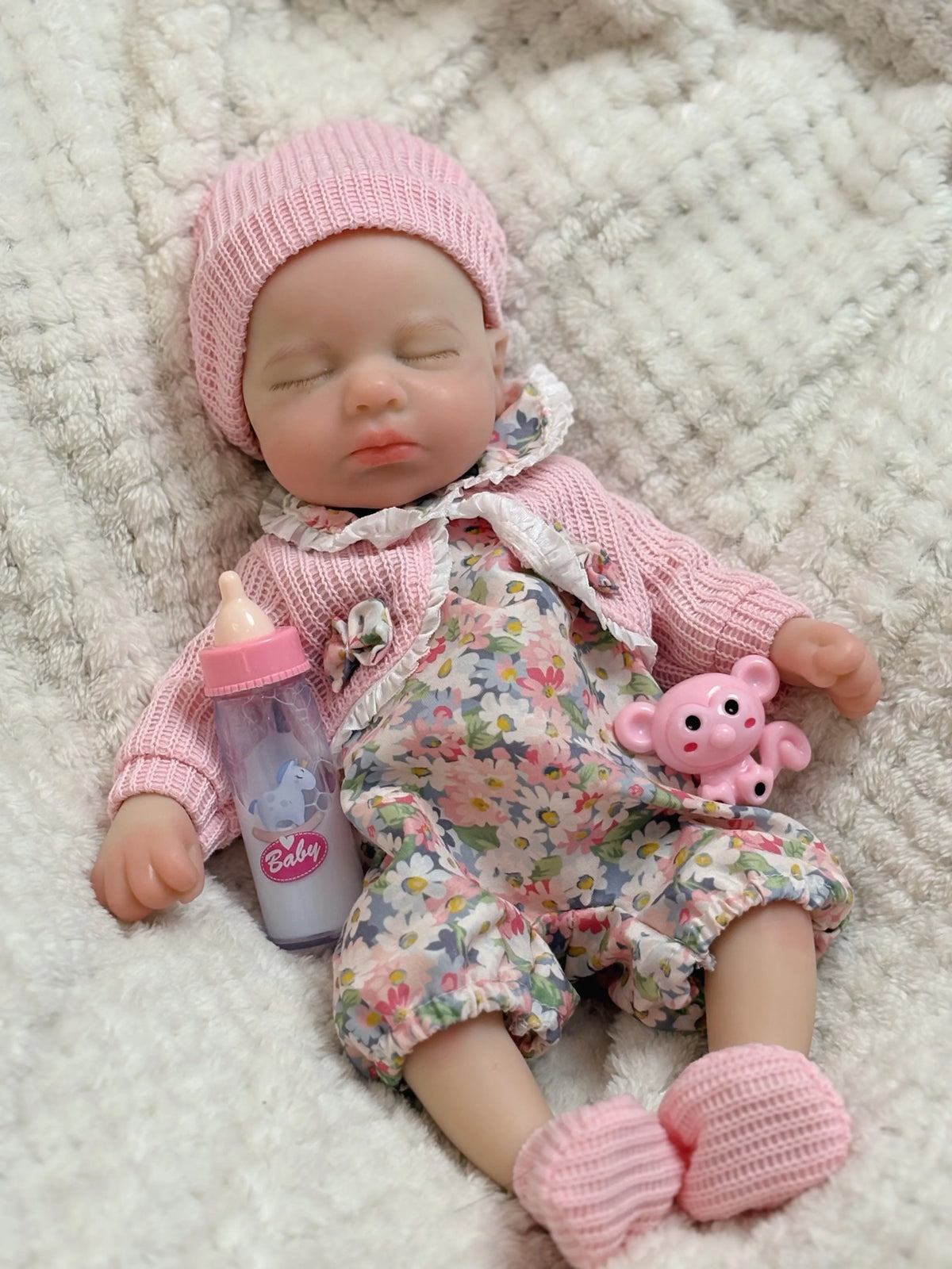 A realistic reborn baby doll wearing a pink knitted hat and floral outfit, lying on a fluffy white surface.