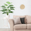 A large green faux monstera palm plant in a white pot sits on a light pink sofa next to a woven wall decoration.