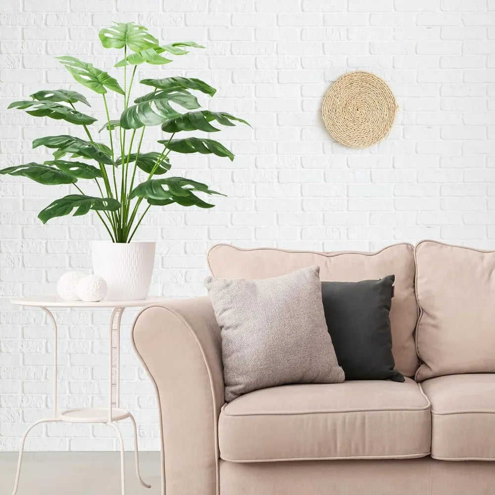 A large green faux monstera palm plant in a white pot sits on a light pink sofa next to a woven wall decoration.