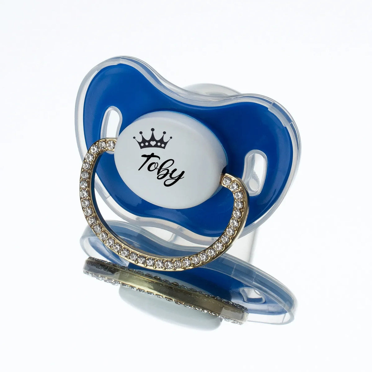 A blue silicone baby pacifier with a white center featuring a gold rhinestone crown and the name Toby.