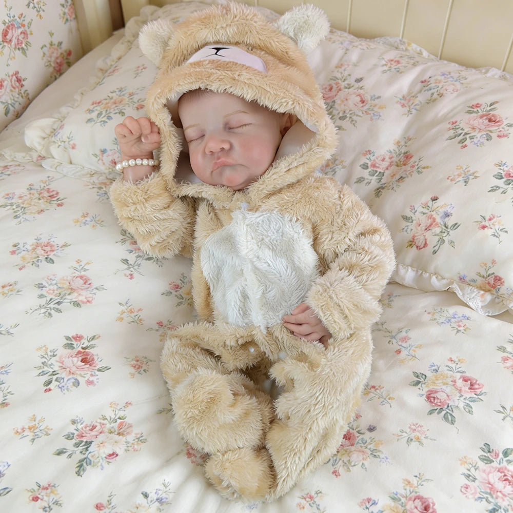 A realistic 18-inch vinyl reborn baby doll wearing a fluffy bear outfit is sleeping on a floral patterned bed.