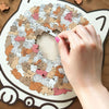 A hand places a pink fingernail on a wooden jigsaw puzzle shaped like a cat, featuring numerous small cats in various colors.