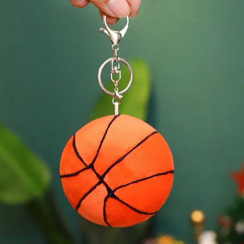 A hand holds a silver sports-themed plush ball with a hook attached, set against a blurred green background.