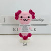 A pink crocheted pencil ornament with purple hair, displayed in front of a white box.