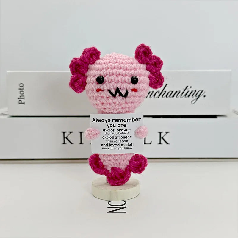 A pink crocheted pencil ornament with purple hair, displayed in front of a white box.