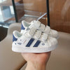 A pair of white and navy casual sneakers with hook-and-loop closure are held in a person's hand.