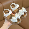 Pair of white sandals with bow details on a tan sole, shown being held by a hand.