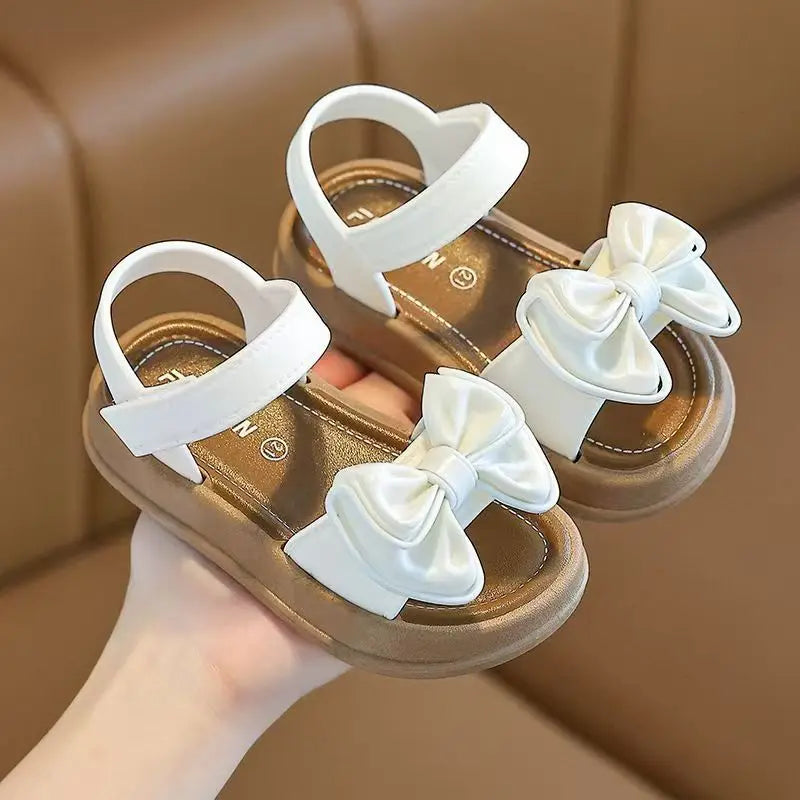 Pair of white sandals with bow details on a tan sole, shown being held by a hand.