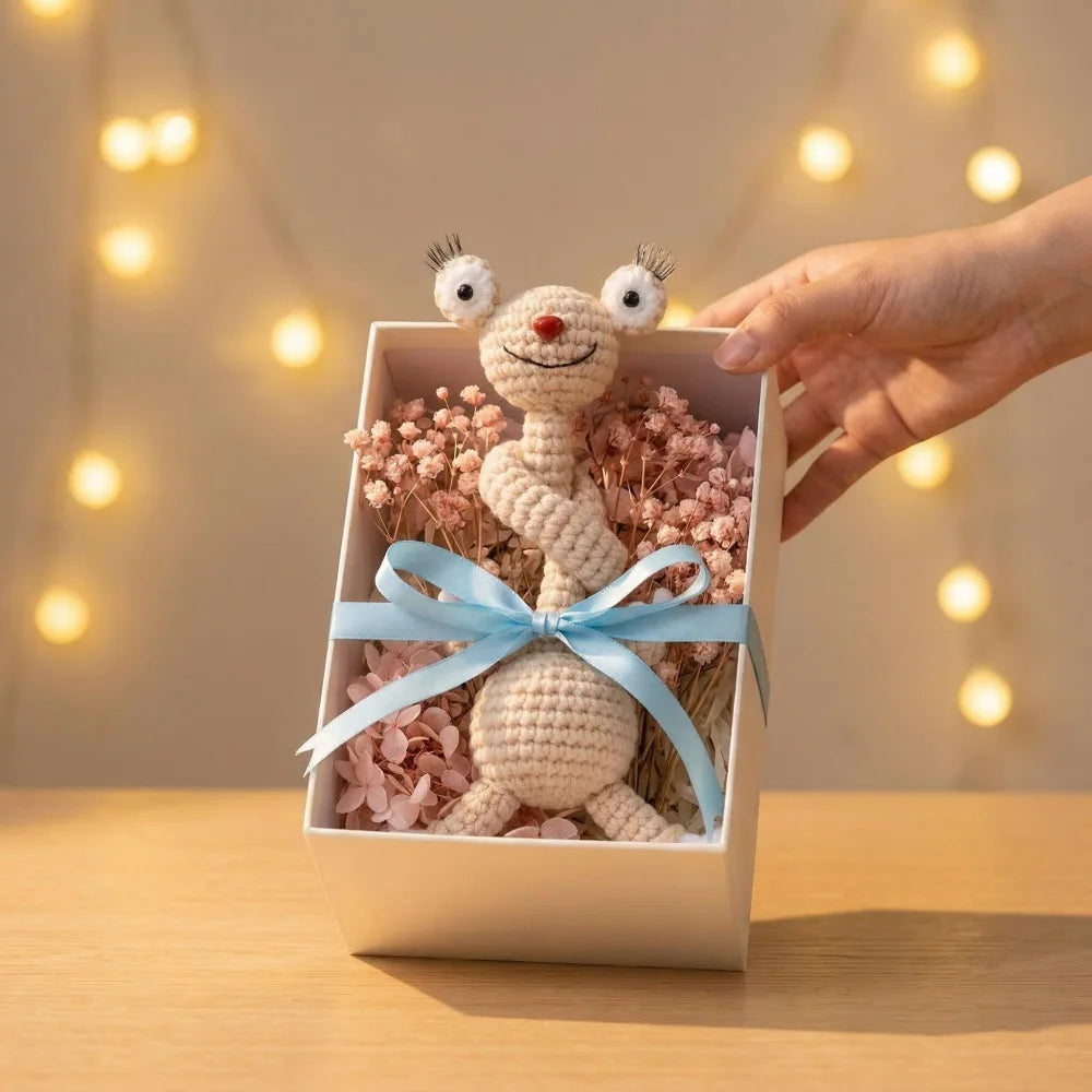A hand places a handmade crochet sloth and squirrel plush charm in a small box surrounded by pink floral elements.