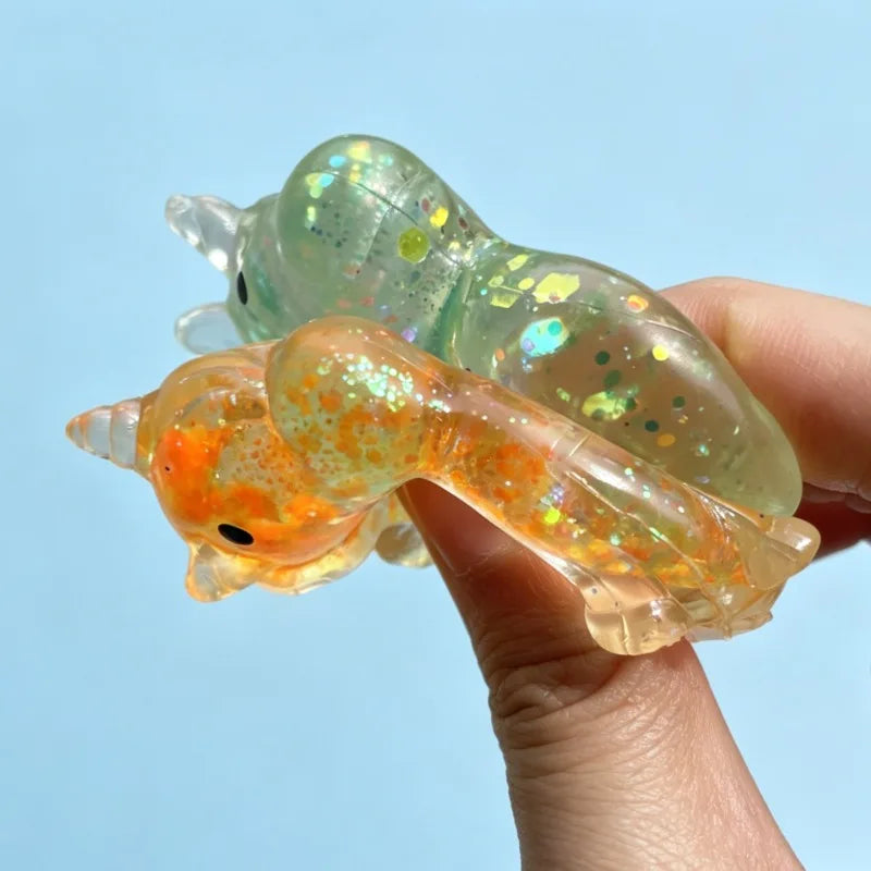 A hand holds a translucent green glitter unicorn stress ball fidget toy against a light blue background.