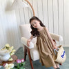 A young girl wearing a beige sleeveless cotton sundress sits in a chair under a modern pendant lamp.