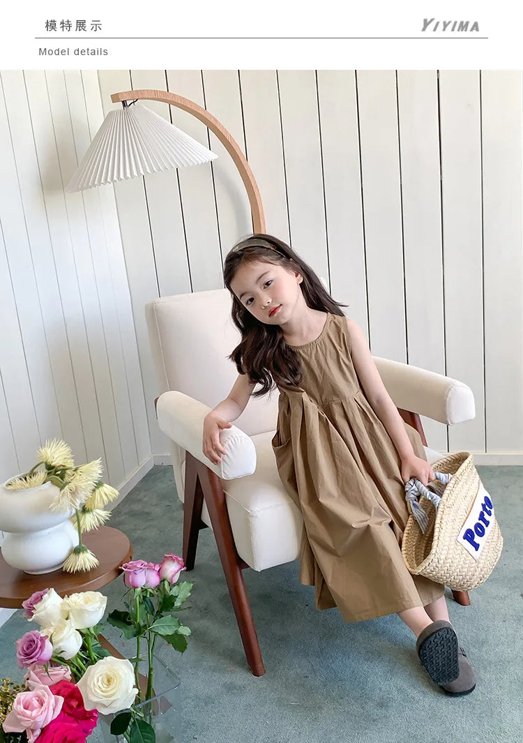 A young girl wearing a beige sleeveless cotton sundress sits in a chair under a modern pendant lamp.